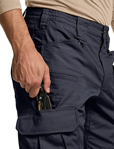Close-up of navy blue tactical cargo pants showing a hand pulling a multi-tool from a side thigh pocket. Multiple pockets visible including a flap-covered cargo pocket below. Dark metal D-ring hardware on waistband.