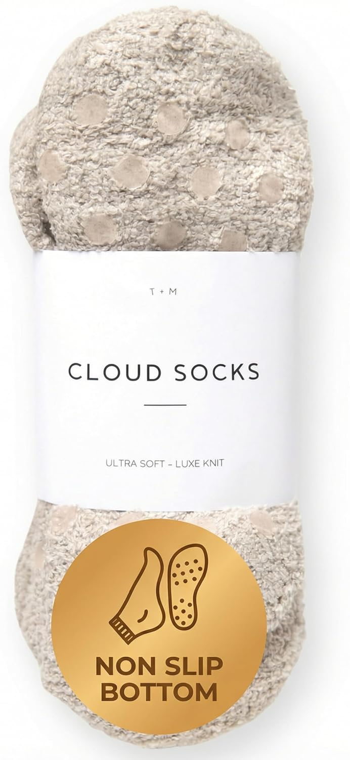 A pair of cream and beige fuzzy socks rolled and banded with a white label reading 'Cloud Socks - Ultra Soft Luxe Knit' by T+M. A gold circular badge at the bottom reads 'Non Slip Bottom' with a sock icon.