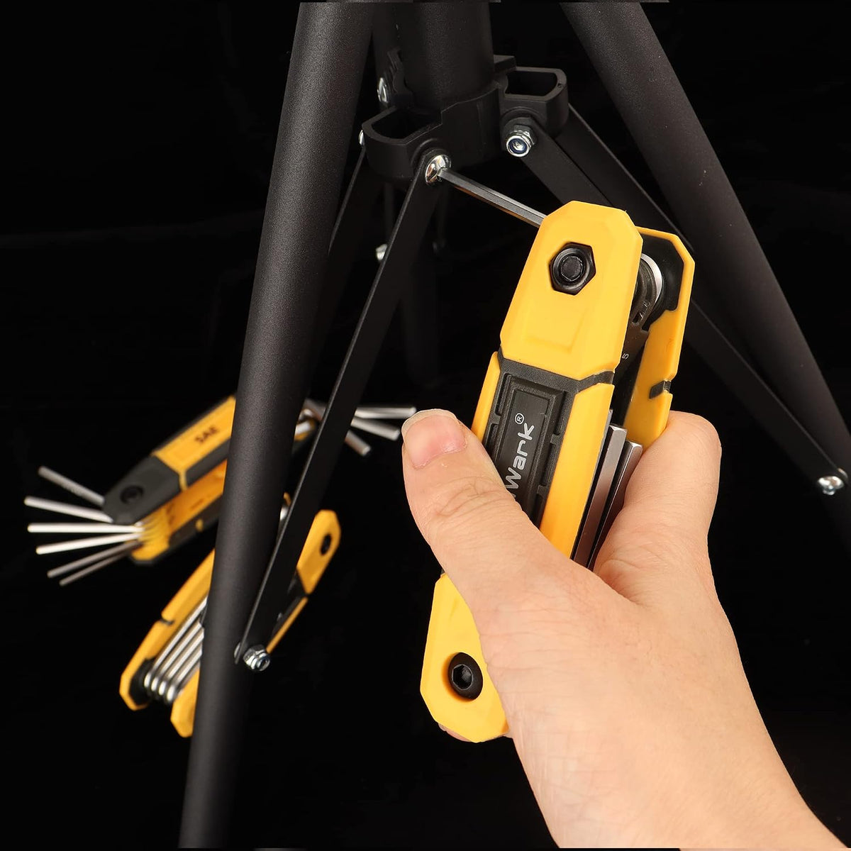 A hand holds a yellow and black folding hex key multi-tool set near a black bicycle frame joint. A second open set with multiple silver hex keys fanned out is visible in the background. Brand label reads 'Wark' on the body.