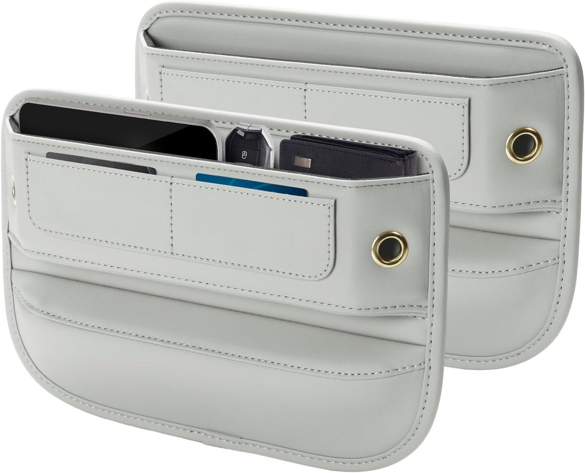 Two light gray leather car seat gap filler organizers shown together, one displaying open pockets holding a phone, card, and other items. Each has stitched compartments, a gold-toned grommet, and curved edges.