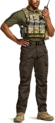 Man wearing dark brown tactical cargo pants with multiple side pockets, paired with an olive green polo shirt, multicam plate carrier vest, and tan boots. Holding a radio, shown from front at full length.