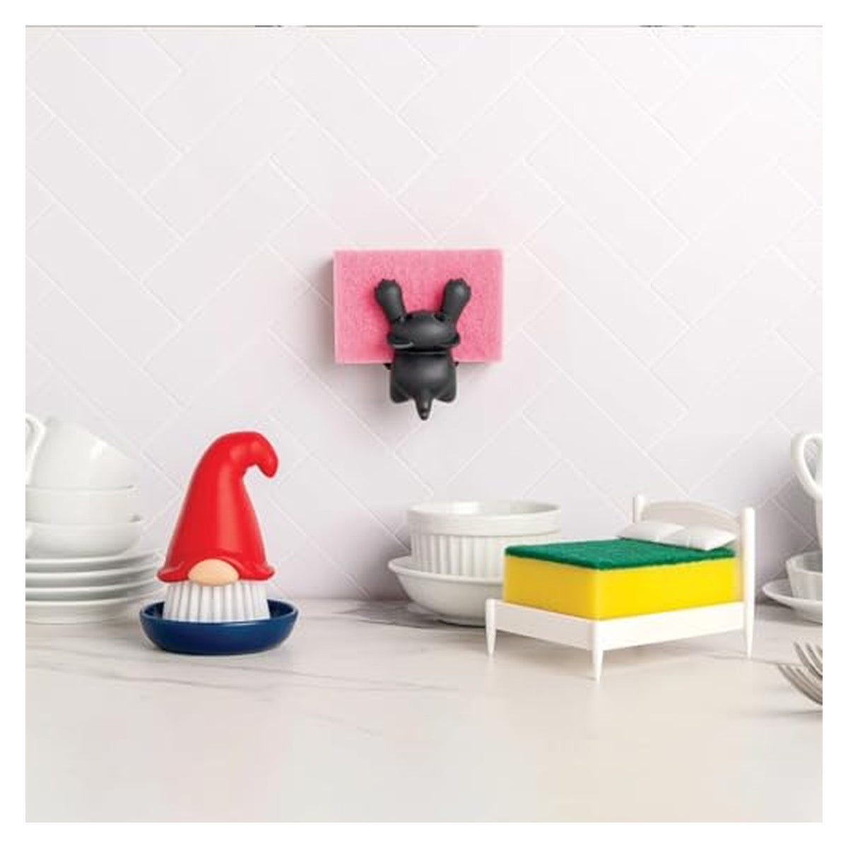 Novelty kitchen accessories on a marble countertop: a gnome-shaped dish brush with red hat and white bristle beard in a blue base, a miniature bed-shaped sponge holder, and a wall-mounted black bunny sponge holder, with white dishes in background.