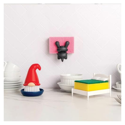 Novelty kitchen accessories on a marble countertop: a gnome-shaped dish brush with red hat and white bristle beard in a blue base, a miniature bed-shaped sponge holder, and a wall-mounted black bunny sponge holder, with white dishes in background.