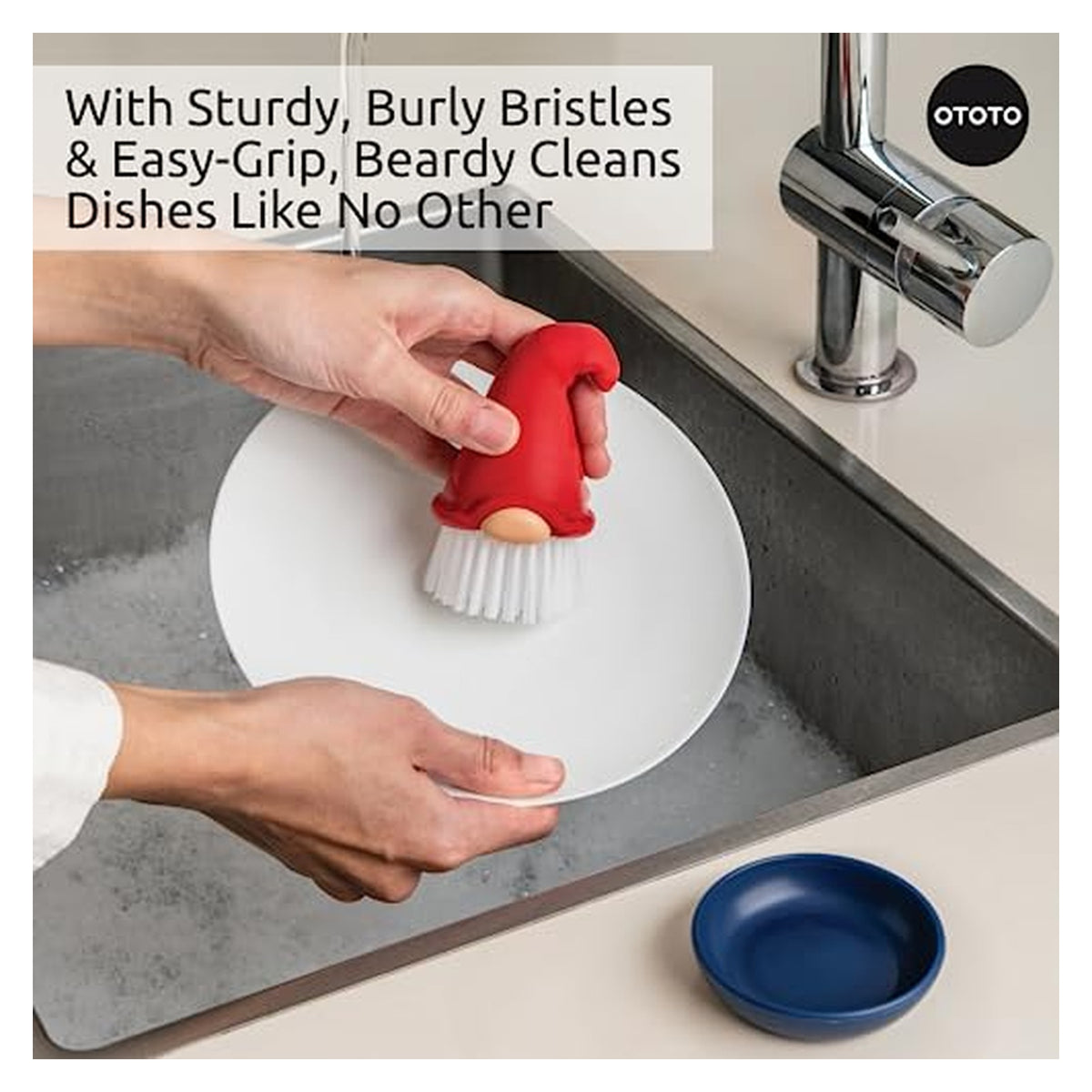 Hands scrubbing a white plate in a sink using a red gnome-shaped dish brush with white bristles and a tan face detail. OTOTO branded. Text reads 'With Sturdy, Burly Bristles & Easy-Grip, Beardy Cleans Dishes Like No Other'