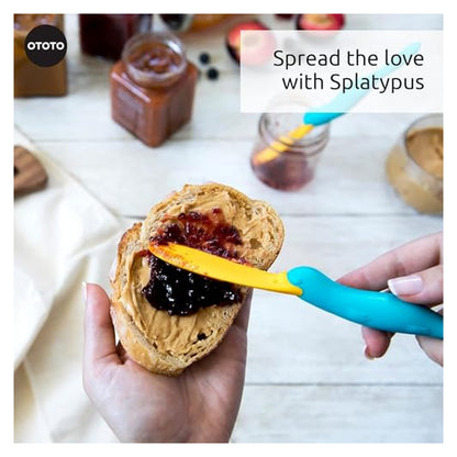 OTOTO Splatypus spreader being used to spread jam on a peanut butter bagel half. The tool has a teal duck-shaped handle and a flat yellow blade. Text reads 'Spread the love with Splatypus.'