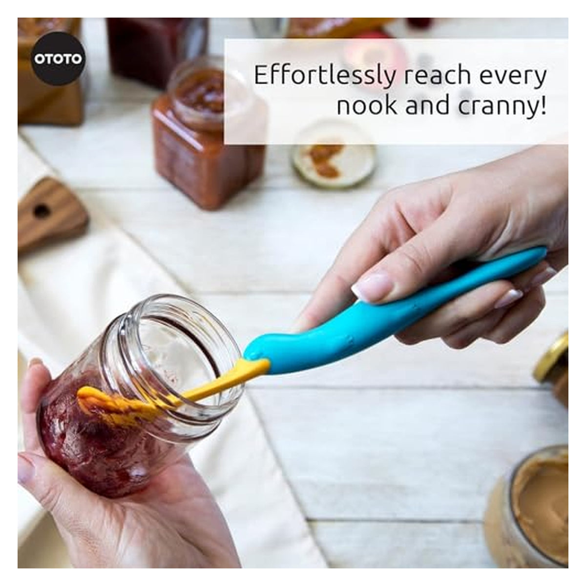 Two hands using the OTOTO Splatypus jar spatula to scrape inside a glass jar of jam. The spatula has a teal handle and a flexible yellow angled tip. Text reads 'Effortlessly reach every nook and cranny!'