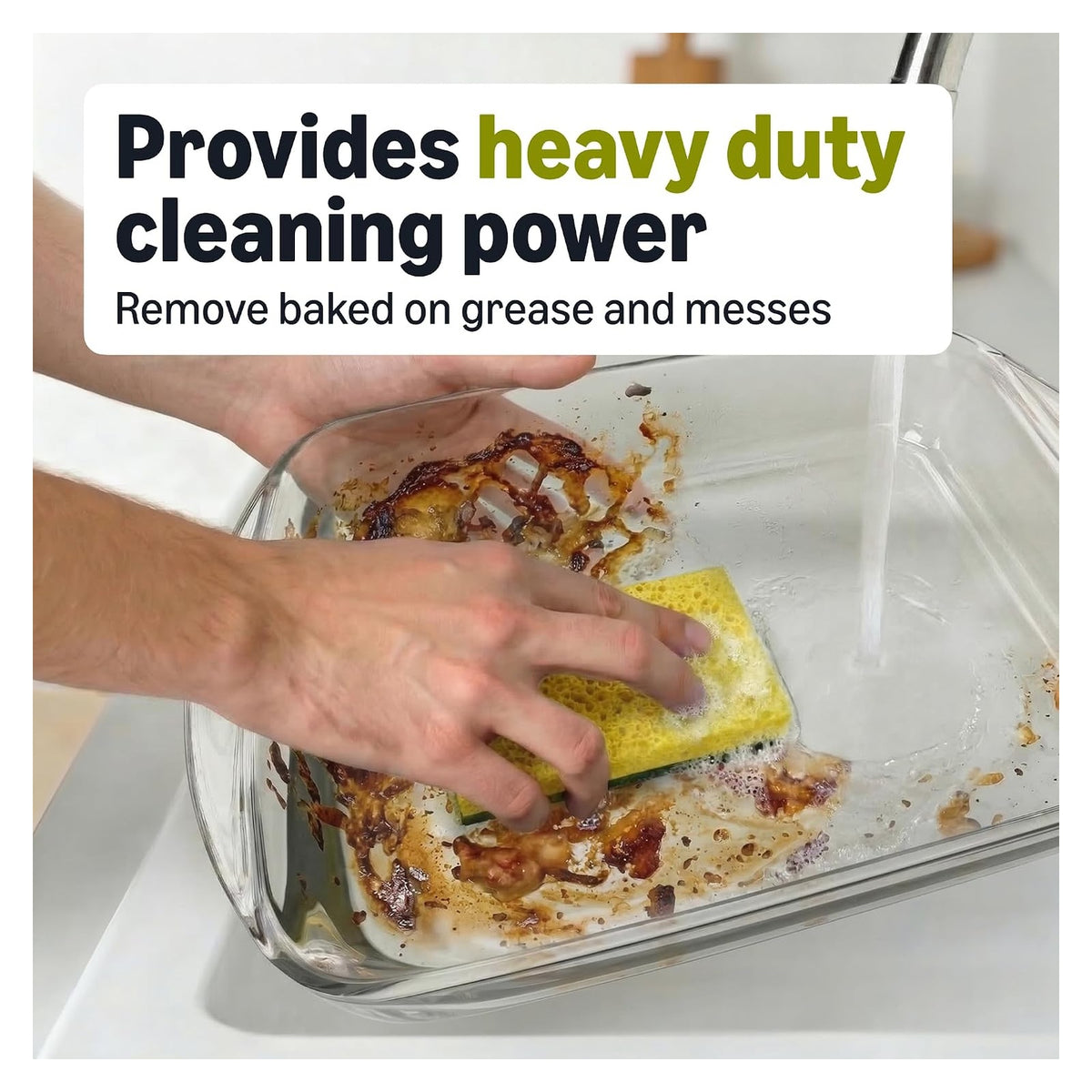 Hands scrubbing a heavily soiled glass baking dish with a yellow-green dual-sided sponge under running water. Text reads 'Provides heavy duty cleaning power - Remove baked on grease and messes.'