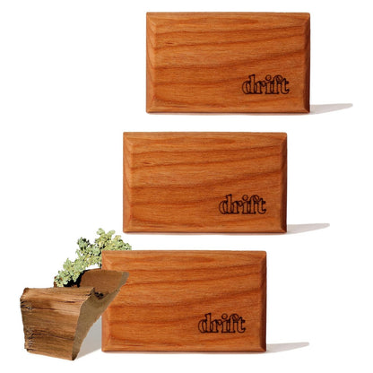Three rectangular wood air fresheners with warm medium-brown finish and visible wood grain, each laser-engraved with the 'drift' logo in the lower right corner. A piece of raw wood with lichen is shown beside the bottom unit.